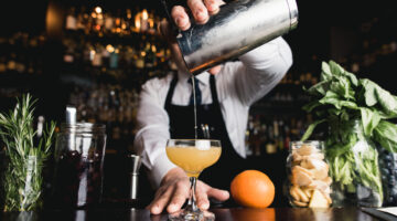 A bar tender pours a cocktail into a glass on a bar