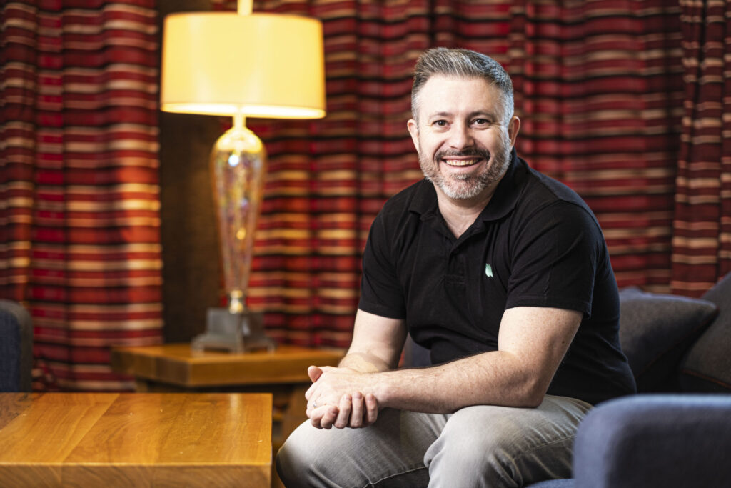 Man, sitting down on blue chair, hands clasped together, wooden table, table lamp, red curtains, Black polo shirt with Nuke from Orbit Logo, grey trousers.