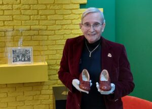 Michael Shaw holds ruby red slippers in his hands. He is wearing a black top with red blazer. He stands in by the corner where a yellow brick wall and green wall meet. There are pictures in a display cabinet behind him.