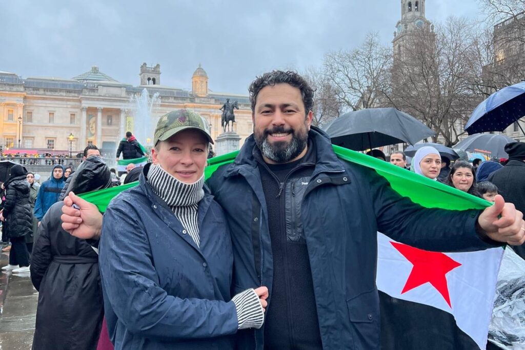 A man and a woman stand holding a Syrian flag in a crowded square.