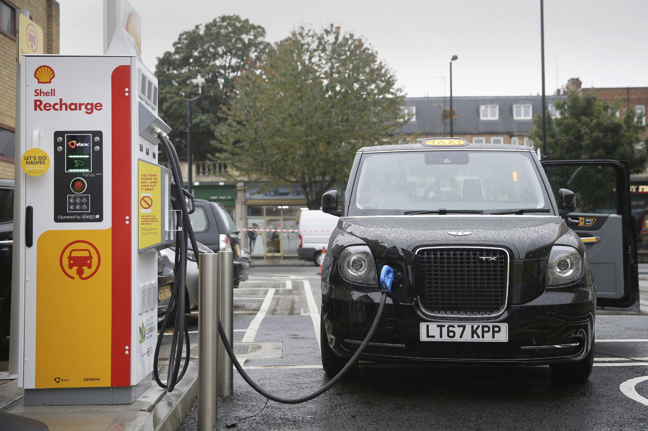A black London taxi plugged into an electric vehicle charger at a Shell Recharge station.